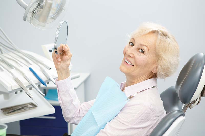 Женщина в возрасте сидит в стоматологическом кресле, смотрит в зеркало на новую улыбку благодаря одномоментной имплантации