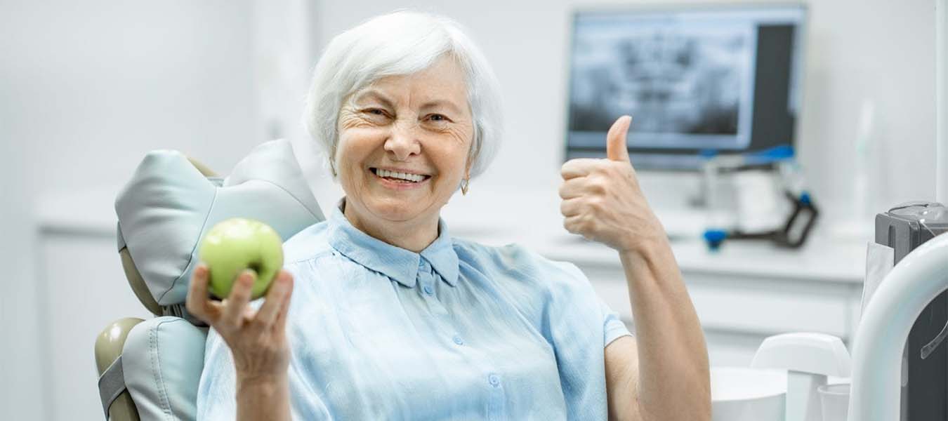 Пожилая женщина держит яблоко и показывает большой палец вверх, что довольна новым крепким и красивым зубам